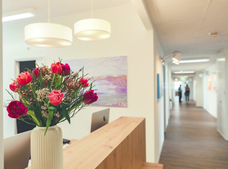 Eine Vase mit roten und rosa Blumen steht auf einem hölzernen Empfangstresen in einem hellen, modernen Büroflur und spiegelt die einladende Atmosphäre einer Praxis für integrative Medizin wider. Zwei Personen gehen den Flur entlang.