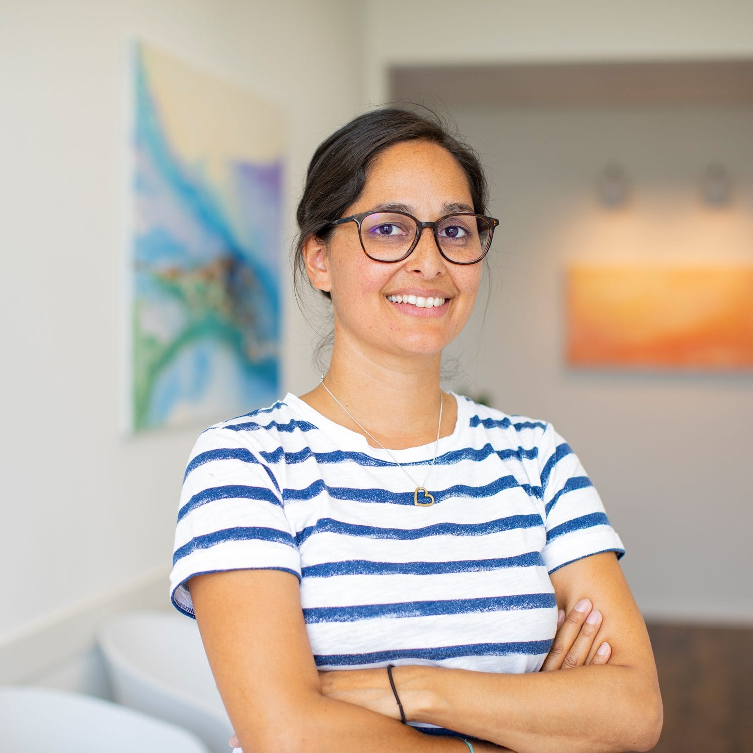 Eine Frau mit Brille und gestreiftem Hemd steht lächelnd mit verschränkten Armen im Raum des Staatsvereins. Hinter ihr hängen farbenfrohe abstrakte Gemälde an den Wänden in einem hellen, modernen Raum.