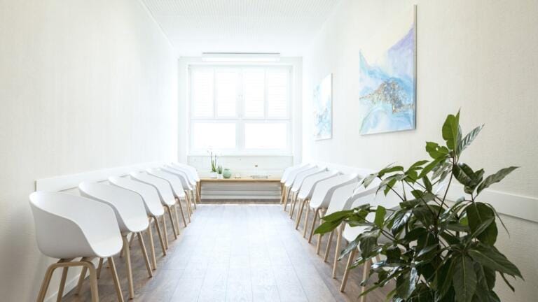 Ein heller, minimalistischer Warteraum im Wise Medicine Gesundheitszentrum verfügt über weiße Wände, moderne weiße Stühle mit Holzbeinen, ein großes Fenster, zwei abstrakte Gemälde und eine grüne Topfpflanze im Vordergrund.