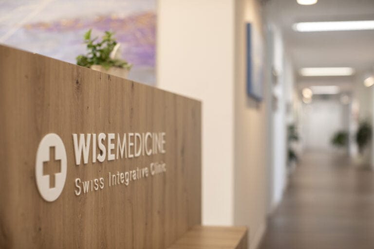 Empfangsbereich der Wise Medicine Swiss Integrative Clinic in Winterthur mit einem Holzschreibtisch, auf dem das Logo der Klinik in weißen Buchstaben prangt; im Hintergrund ist ein sanft beleuchteter Flur zu sehen.