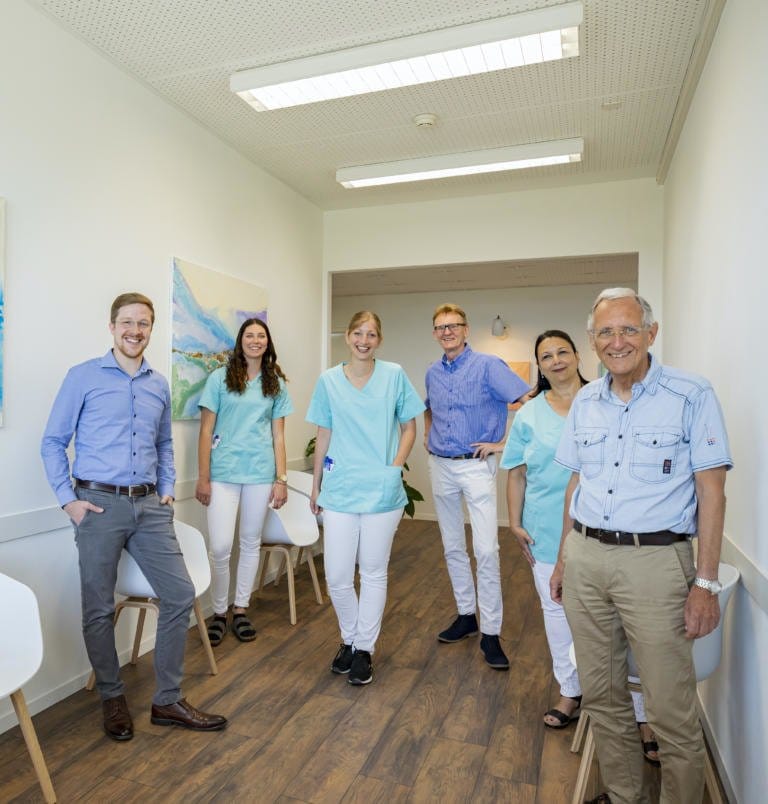 Eine Gruppe von sechs lächelnden Erwachsenen, darunter drei in blauer Arztkleidung und drei in legerer Geschäftskleidung, steht zusammen in einem hellen Flur mit Holzboden und moderner Einrichtung im Wise Medicine Tag der offenen Tür.