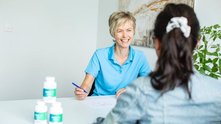 Eine lächelnde medizinische Fachkraft in einem blauen Hemd und mit einem Stift in der Hand berät einen Patienten an einem Schreibtisch mit Body Recharge-Nahrungsergänzungsmittelflaschen und Papieren in Claudia Kaisers hellem Büro.