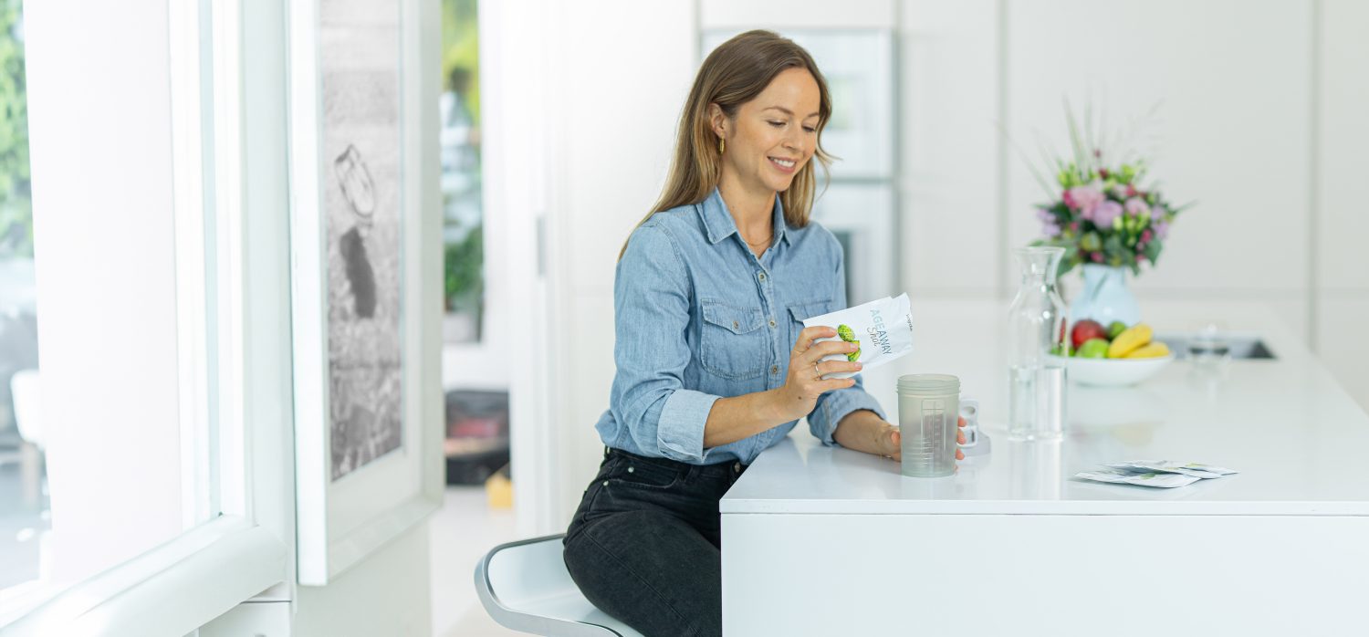 Eine Frau sitzt an einem weißen Küchentisch und schüttet lächelnd die Nahrungsergänzung aus der Schweiz aus einer Packung in eine Shakerflasche. Ein Glas Wasser, eine Schale mit Obst und eine Vase mit Blumen stehen auf dem Tresen daneben.