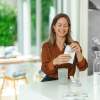 Eine lächelnde Frau sitzt an einer hellen Küchentheke und öffnet eine Packung „AG1“ von kingnature Nahrungsergänzung aus der Schweiz neben einer Glaskaraffe mit Wasser, einer Shaker-Flasche und anderen Packungen mit Nahrungsergänzungsmitteln.