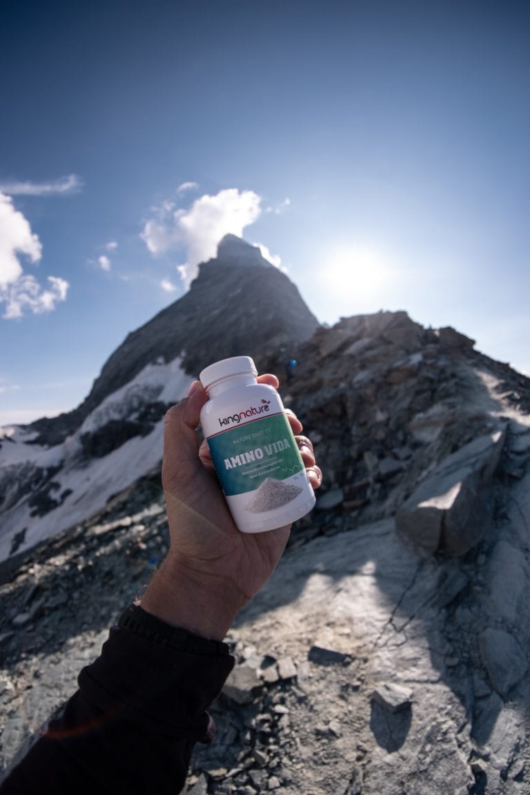 Jemand hält eine Flasche Amino Vida Aminosäuren Kapseln mit dem Etikett vor einem Berggipfel und strahlender Sonne unter einem klaren blauen Himmel.