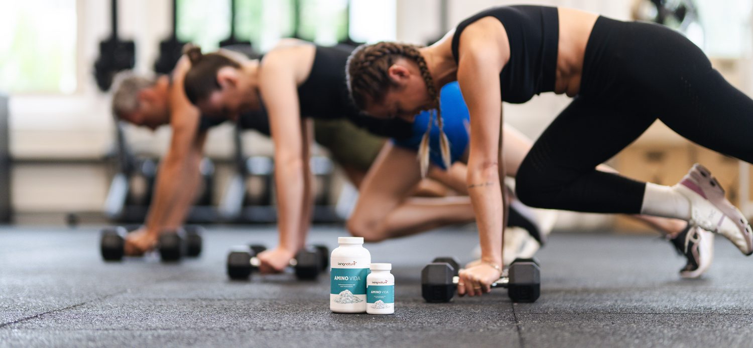Three people are working out with dumbbells in a gym, blurred in the background. In the foreground are three bottles of Amino Lion by kingnature, a premium dietary supplement from Switzerland. The setting appears to be a gym.