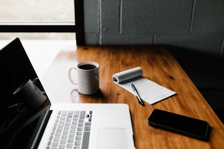 Ein Holzschreibtisch mit einem aufgeklappten Laptop, einer Tasse Kaffee, einem Stift auf einem Notizblock mit handschriftlichen Notizen zum Thema Vitalstoffmedizin und einem Smartphone. Sonnenlicht fällt durch ein nahegelegenes Fenster – perfekt für die Teilnahme an den Kostenlosen Webinaren.