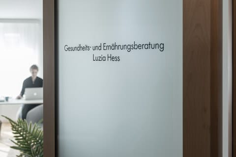 Milchglastür mit dem Schriftzug Gesundheits- und Ernährungsberatung Luzia Hess. Im Hintergrund sitzt eine Person mit Laptop an einem Schreibtisch im hellen Büro der Luzia Hess GmbH.