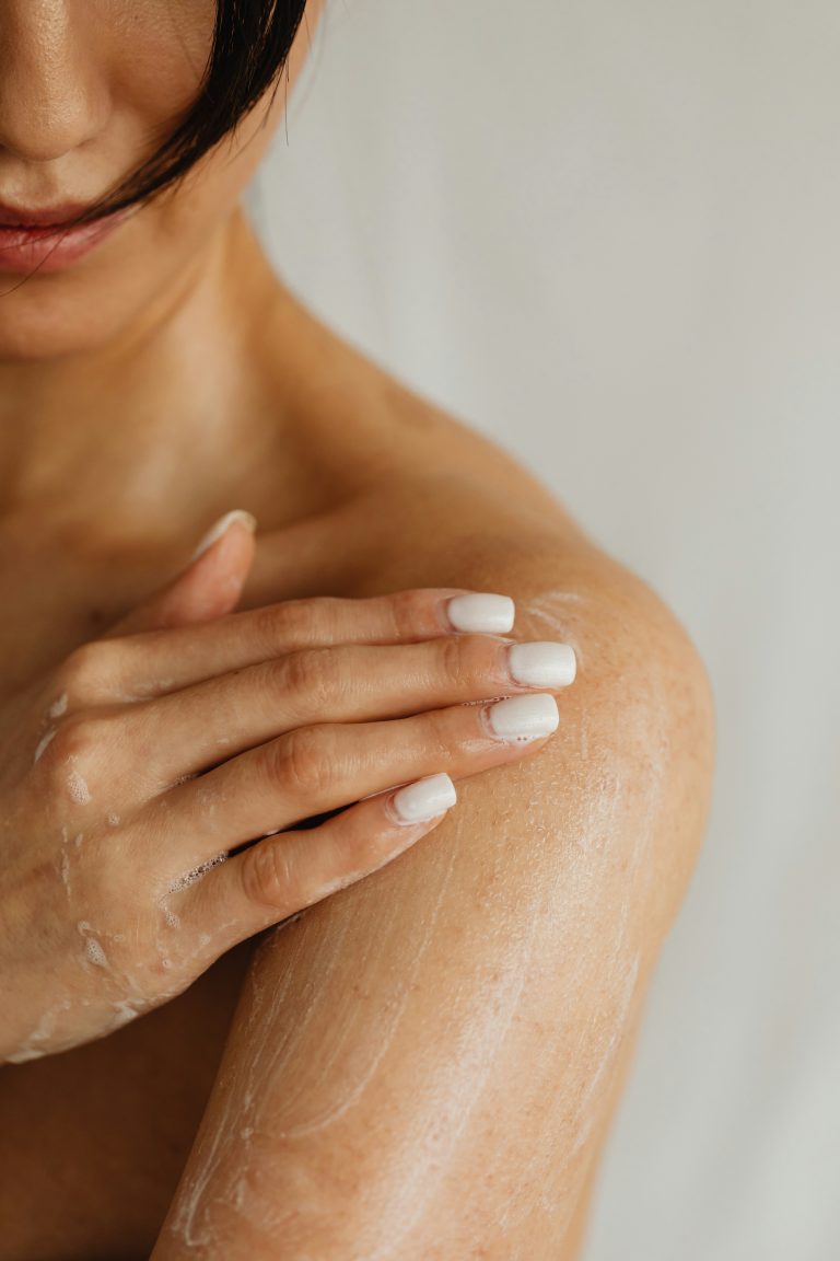 Eine Frau mit heller Haut trägt Creme auf ihre nackte Schulter auf und zeigt dabei vor einem weichen, neutralen Hintergrund gepflegte weiße Nägel und ein teilweise verdecktes Gesicht – was auf die Rolle von Biotin für gesunde Haut und Nägel hindeutet.
