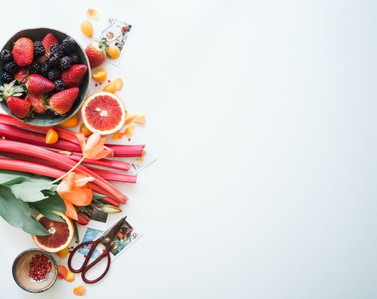 Ein farbenfrohes Arrangement aus Erdbeeren, Brombeeren, Kumquats, Blutorangenscheiben und Rhabarber auf einer weißen Oberfläche mit Blumen und Samenpaketen hebt lebenswichtige Nährstoffe für den Körper hervor und lässt auf der rechten Seite viel Platz frei.