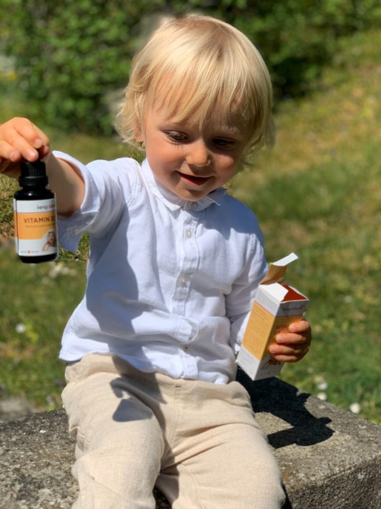 Ein kleines Baby mit blondem Haar, bekleidet mit einem weißen Hemd und einer hellen Hose, sitzt im Freien, hält in der einen Hand eine Flasche Vitamin-D3-Babytropfen und in der anderen die Produktverpackung und lächelt strahlend im Sonnenlicht.