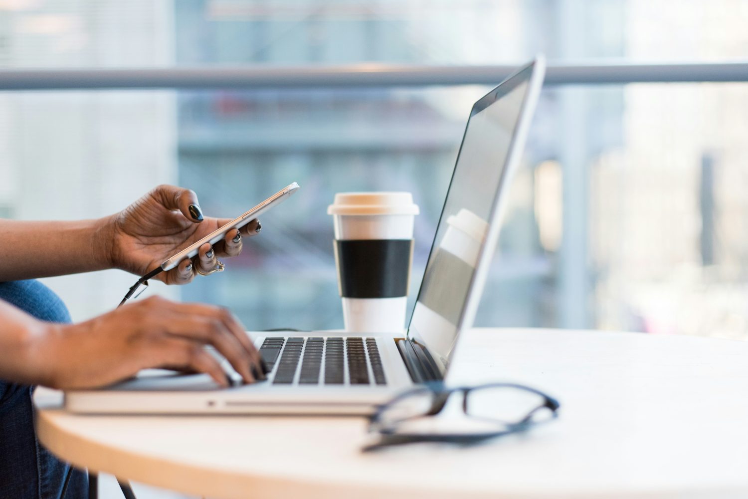 Eine Person, die an einem Tisch einen Laptop benutzt und ein Smartphone in der Hand hält, mit einer Kaffeetasse zum Mitnehmen, einer Brille und Mitoflow Vida - dem neuen Produkt zur Unterstützung von Mitochondrien - in der Nähe. Der Hintergrund zeigt eine helle, moderne Innenumgebung mit großen Fenstern.