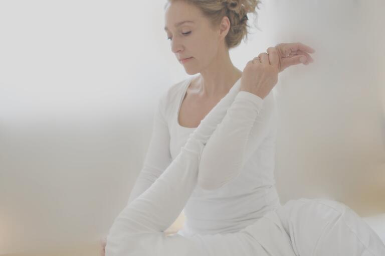 Anna Goetsch übt in weißer Kleidung eine Yoga-Pose, sitzt ruhig da und streckt ihren Arm über ihren Kopf in einem hellen, sanft beleuchteten Raum der Shiatsu-Praxis.