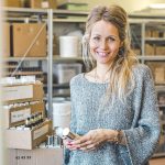 Eine lächelnde Frau mit langen blonden Haaren und einem grauen Pullover steht in einem Lagerraum und hält kleine Flaschen in der Hand. Im Hintergrund sind Kisten und Regale mit verschiedenen Naturprodukten zu sehen.