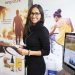 Eine lächelnde Frau mit langen braunen Haaren und Brille steht drinnen und hält Notizblock und Stift in der Hand. Sie trägt ein schwarzes Kleid und steht neben einem Schreibtisch. Werbebanner mit Nahrungsergänzungsmitteln und einer sportlichen Frau, die für Wellness wirbt.