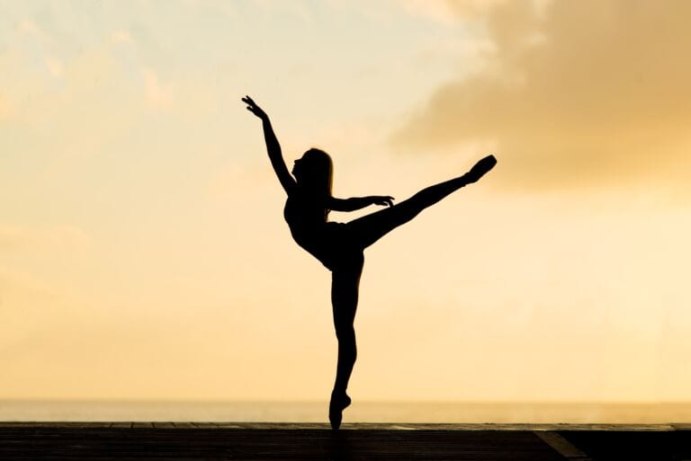 Silhouette einer Balletttänzerin, die auf einem Fuß balanciert, wobei sie ein Bein weit nach hinten ausgestreckt und die Arme anmutig erhoben hat, vor dem Hintergrund eines warmen, goldenen Sonnenuntergangshimmels – eine Szene, die dazu inspiriert, Bindegewebe zu stärken, möglicherweise unterstützt durch Nahrungsergänzung.