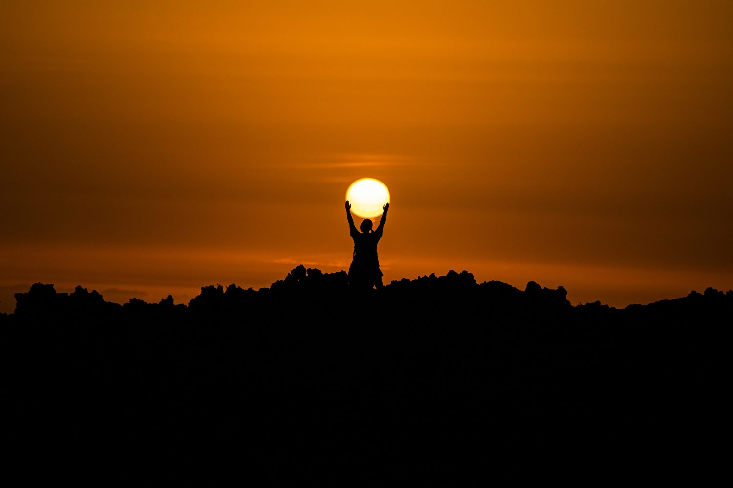 Die Silhouette einer Person mit „Sonnenverstand“ steht auf felsigem Boden und scheint die untergehende Sonne vor einem orangefarbenen Himmel bei Sonnenuntergang emporzuhalten.