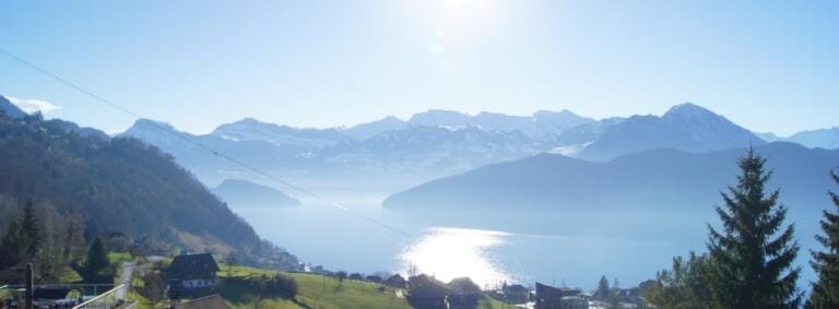 Ein wunderschöner, heller, sonniger Tag mit Blick auf einen ruhigen See, umgeben von Bergen, mit grünen Hügeln, verstreuten Häusern und Kiefern im Vordergrund. Sonnenlicht schimmert auf dem Wasser unter einem klaren blauen Himmel.