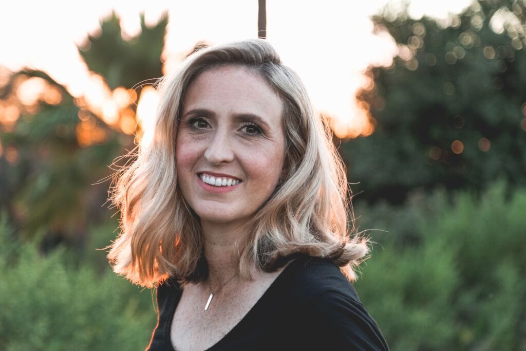 Eine Frau mit blondem, welligem Haar und einem schwarzen Oberteil lächelt im Freien bei Sonnenuntergang, umgeben von grünem Laub – und genießt die Unterstützung während ihrer Wechseljahre.