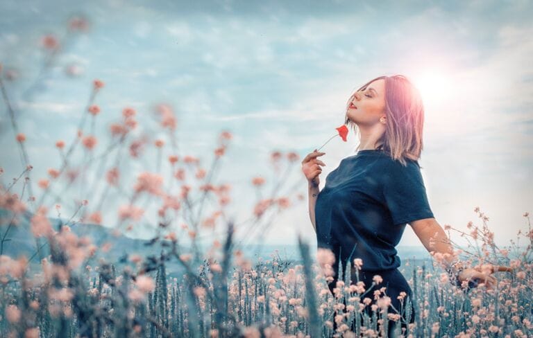 Eine Frau steht in einem Feld voller Wildblumen, hat die Augen geschlossen und hält eine rote Blume vor ihr Gesicht. Hinter ihr leuchtet das Sonnenlicht, und über ihr ist ein sanfter, pastellfarbener Himmel zu sehen – ein Zeichen für die sanfte Unterstützung während der Wechseljahre.