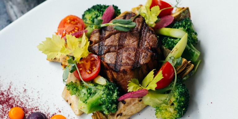 Gegrilltes Steak serviert mit Brokkoli, Kirschtomaten, Sellerieblättern und anderem farbenfrohen Gemüse, kunstvoll auf einem weißen Teller angerichtet für eine köstliche und farbenfrohe Mahlzeit.