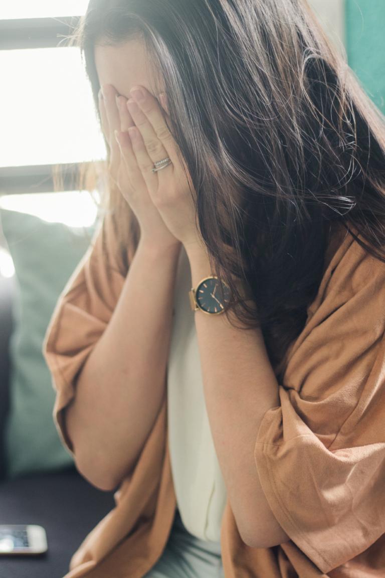 Eine Frau mit langen braunen Haaren, einer beigen Strickjacke und einer Armbanduhr sitzt auf einem Sofa, die Hände vor dem Gesicht, und scheint von Schmerz erfasst zu sein. Ein Smartphone liegt daneben, während sie über die Bedeutung von Schmerz nachdenkt.
