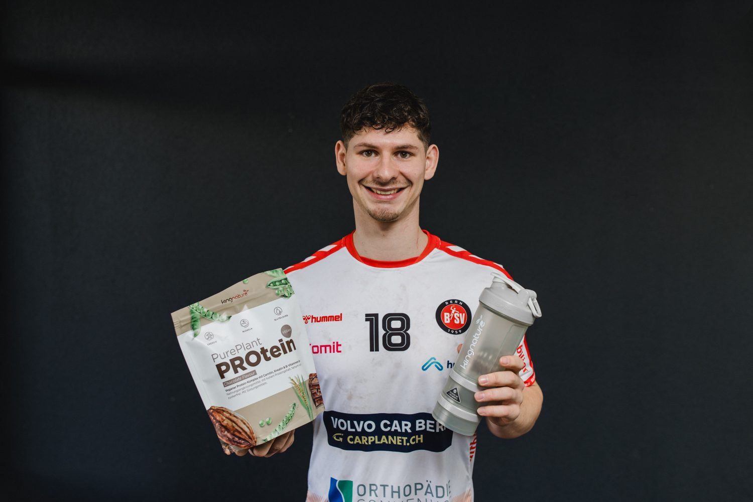 A smiling man in a sports jersey holds a bag of PurePlant Protein powder in one hand and a shaker bottle in the other, standing against a plain dark background.