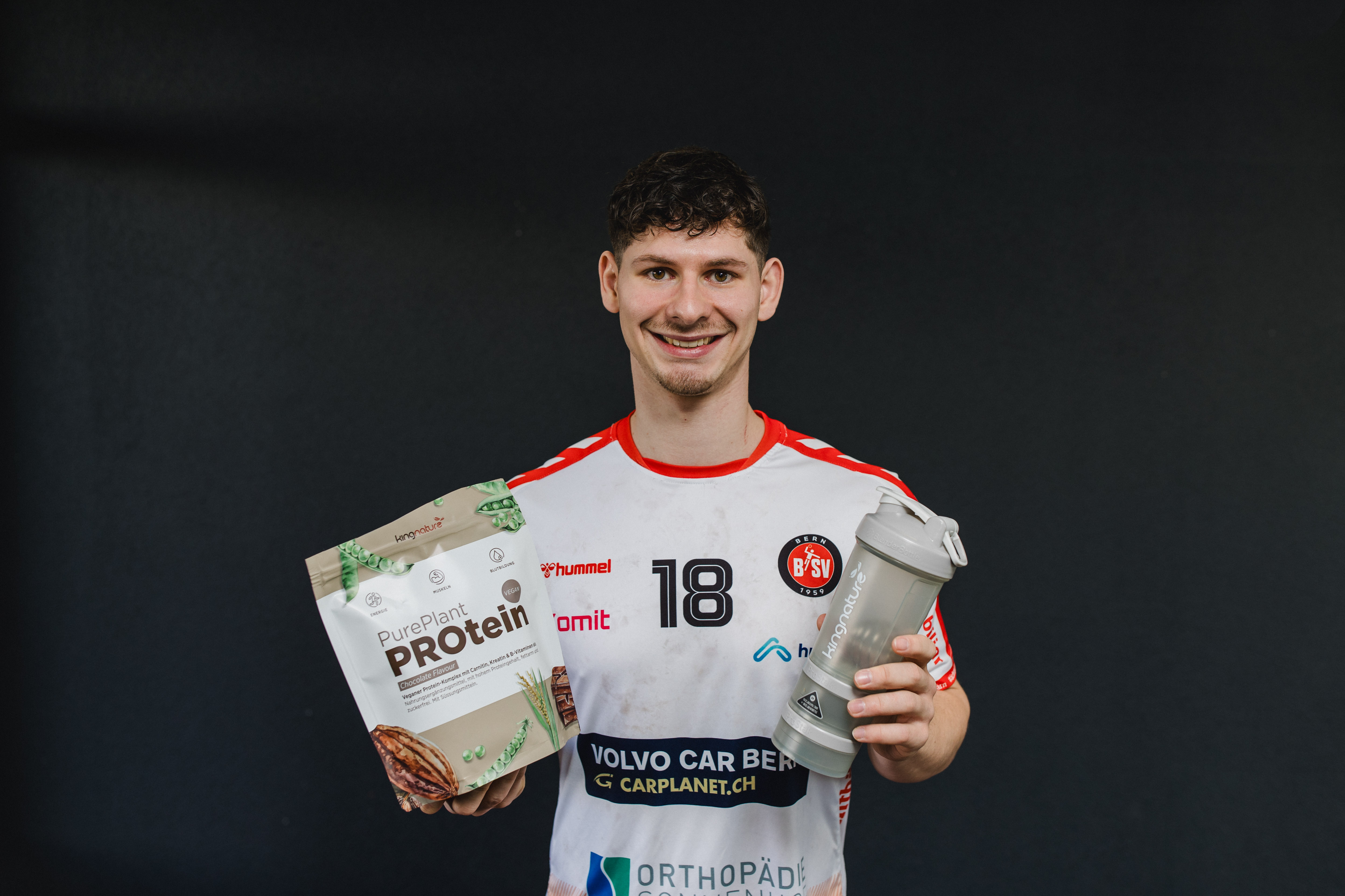 A smiling man in a sports jersey holds a bag of PurePlant Protein powder in one hand and a shaker bottle in the other, standing against a plain dark background.