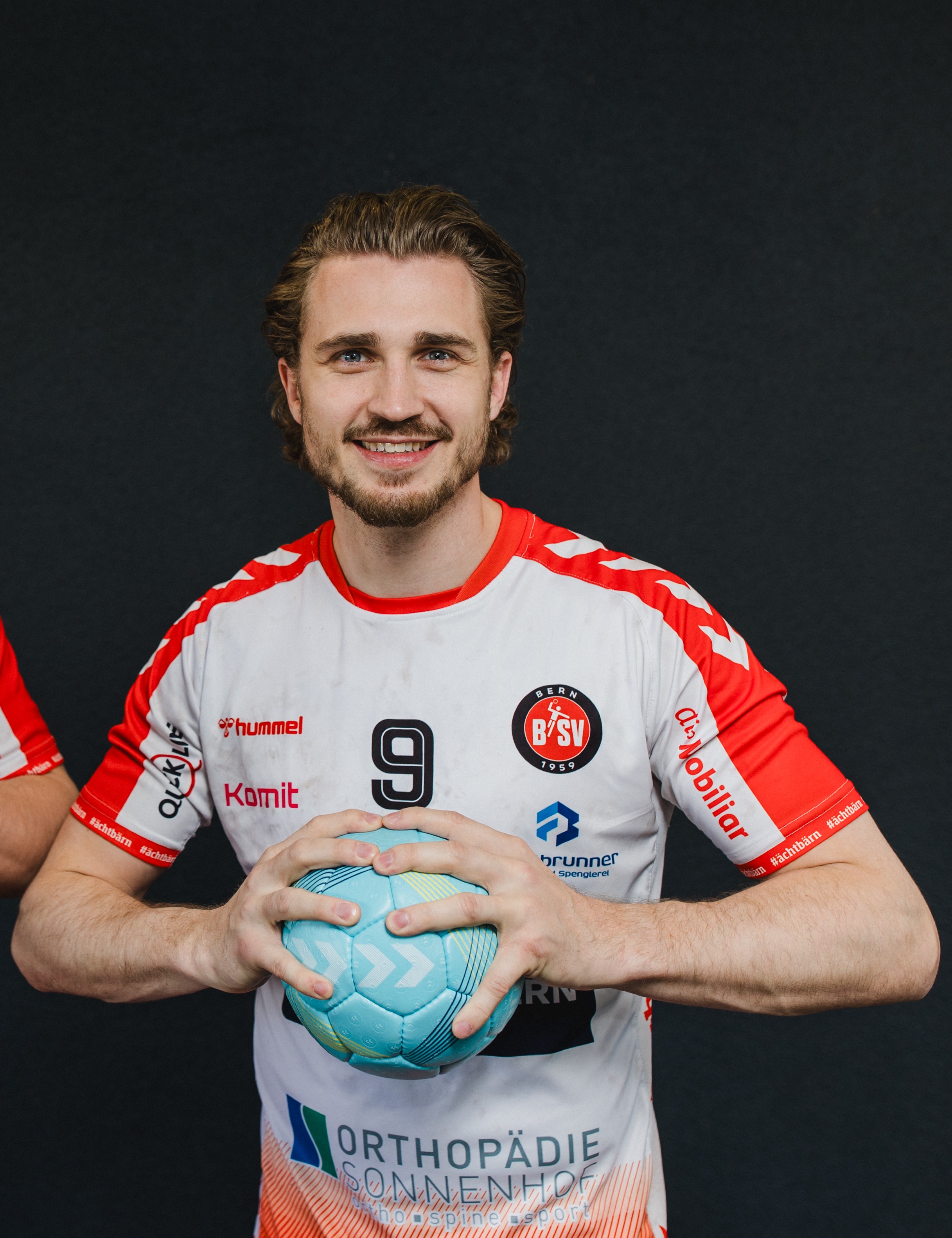 Ein lächelnder männlicher Handballspieler in einem weiß-roten Trikot mit der Nummer 9 hält einen blauen Ball vor einem dunklen Hintergrund und verkörpert damit die Energie und Kraft, die Amino Vida unterstützen kann.