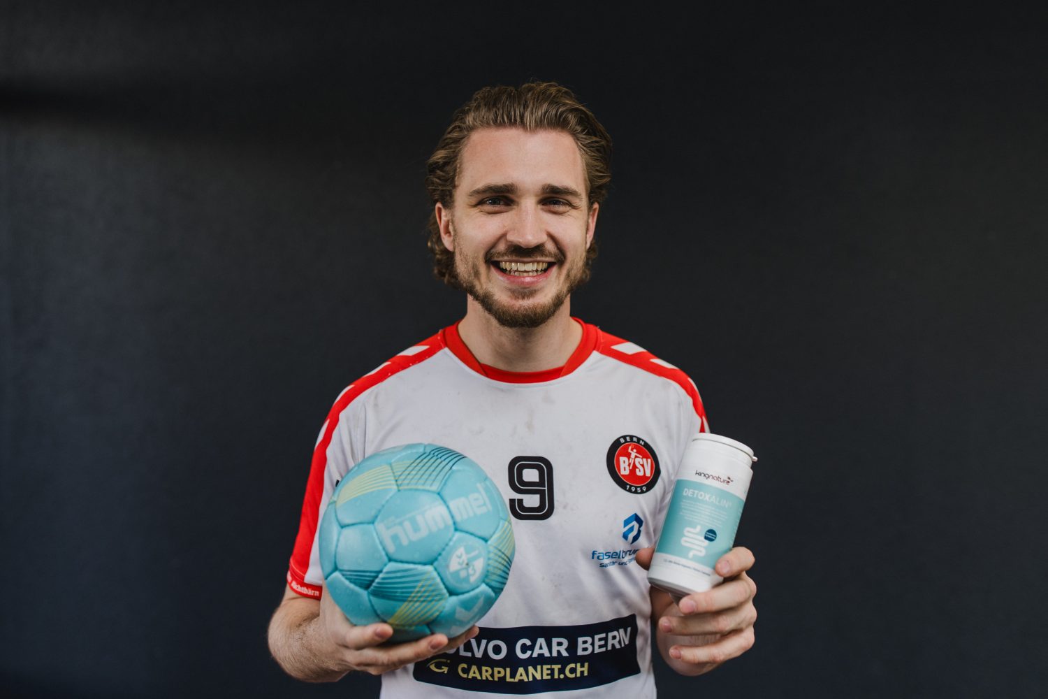 A smiling man in a white and red sports jersey holding a blue soccer ball in one hand and a beverage cup in the other, standing against a dark background.