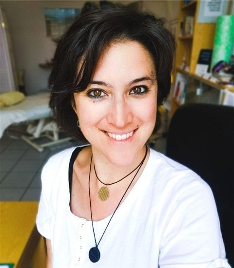 Eine Frau mit kurzen dunklen Haaren und grünen Augen, die ein weißes Hemd und zwei Halsketten trägt, lächelt in die Kamera in einem hell erleuchteten Raum mit Regalen und einem Bett im Hintergrund, was die Wärme der Mountain Anna-Therapie widerspiegelt.