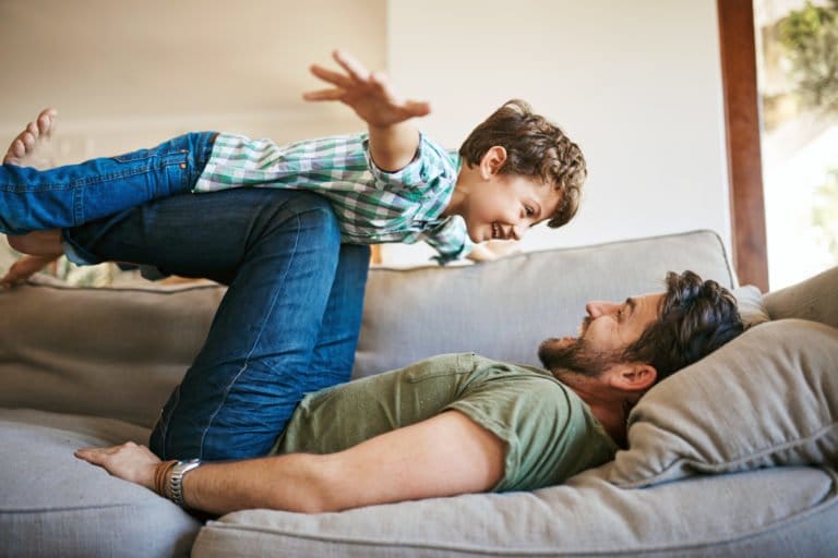 Ein lächelnder Mann liegt auf einer Couch und hebt einen glücklichen Jungen mit seinen Beinen in die Luft. Der Junge streckt seine Arme wie ein Flugzeug aus, und beide lachen – sie genießen die gemeinsame spielerische Zeit nach ihrer ADHS-Beratung.