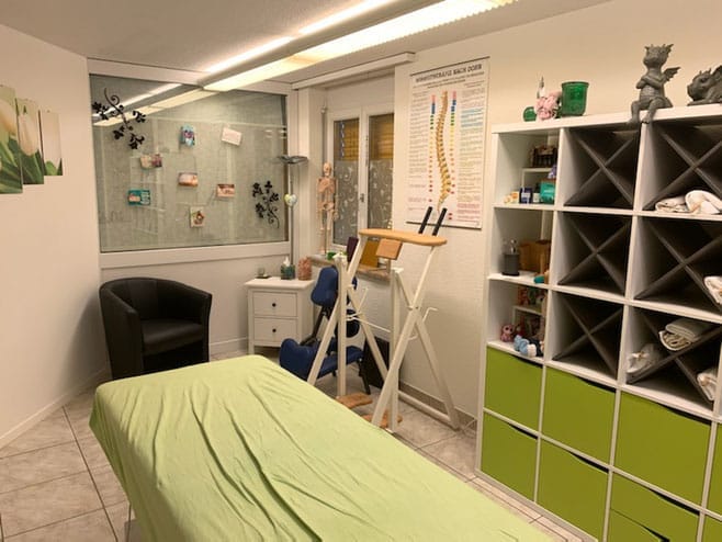 Ein heller Massagetherapieraum für die Myoreflextherapie von Lüthi besticht durch eine grün bezogene Massageliege, einen schwarzen Stuhl, weiße Regale, ein anatomisches Wirbelsäulenposter und ein mit Fotos und Karten geschmücktes Fenster. Der geflieste Boden trägt zum sauberen, übersichtlichen Ambiente bei.