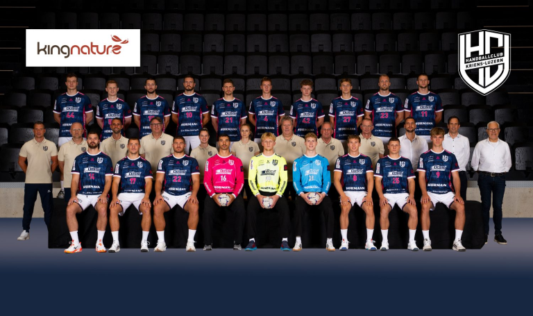 Eine Handballmannschaft posiert für ein Gruppenfoto in einer Halle und strahlt sportliche Freude aus. Die Spielerinnen und Spieler in blauen Trikots und die Betreuerinnen und Betreuer in beigen Hemden sind in drei Reihen aufgestellt, im Hintergrund sind die Logos von Kingnature und des Handballclubs Kriens-Luzern zu sehen.