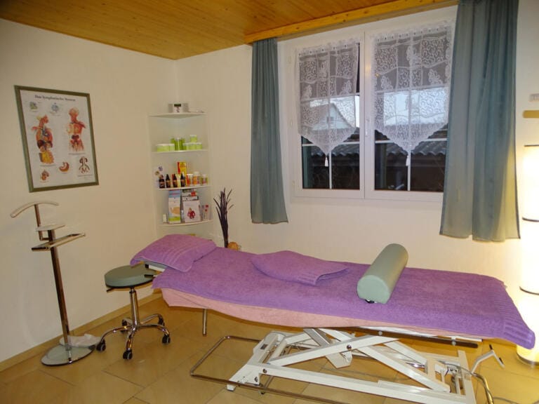 Ein sauberer, gut beleuchteter Behandlungsraum der Praxis verfügt über einen Massagetisch mit lila Handtüchern, einen Rollhocker, medizinisches Zubehör auf Regalen, ein anatomisches Poster an der Wand und ein Fenster mit Spitzenvorhängen – ideal für Wohl im Bauch.