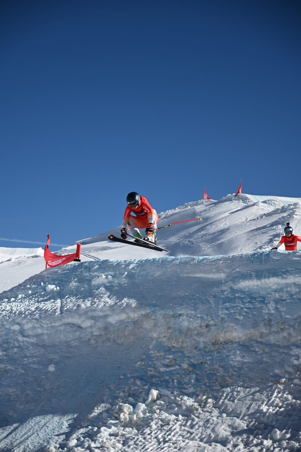A skier in a red outfit and helmet jumps off a snowy slope during a ski race, with red flags marking the course against a clear blue sky and snowy mountain background.