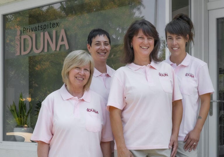 Vier Frauen in passenden hellrosa Poloshirts mit Logos der IDUNA GmbH stehen lächelnd vor einem Gebäude mit einem Schild mit der Aufschrift „Privatspitex IDUNA“. Durch die Glastür sind Pflanzen im Inneren sichtbar.