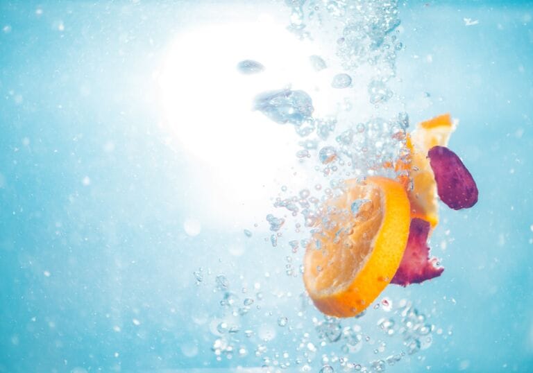 Orangen- und rote Fruchtscheiben plätschern in klares blaues Wasser, umgeben von Blasen und beleuchtet von hellem Sonnenlicht, und symbolisieren Mikronährstoffe, die dein Immunsystem unterstützen.