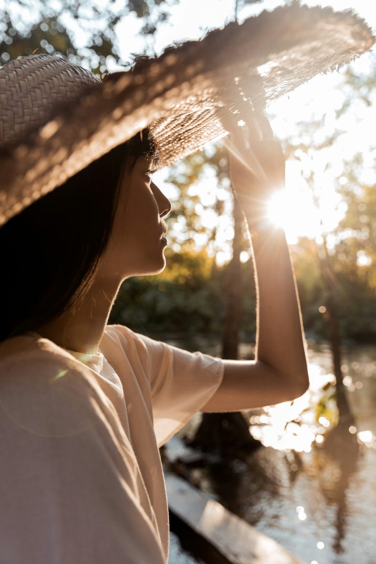 Eine Person in einem weißen Hemd und einem Strohhut hält die Hutkrempe fest, während das Sonnenlicht durch die Bäume am Flussufer scheint – ein Ausdruck von wahrem „Sonnenverstand“, der einen warmen, von hinten beleuchteten Effekt erzeugt.