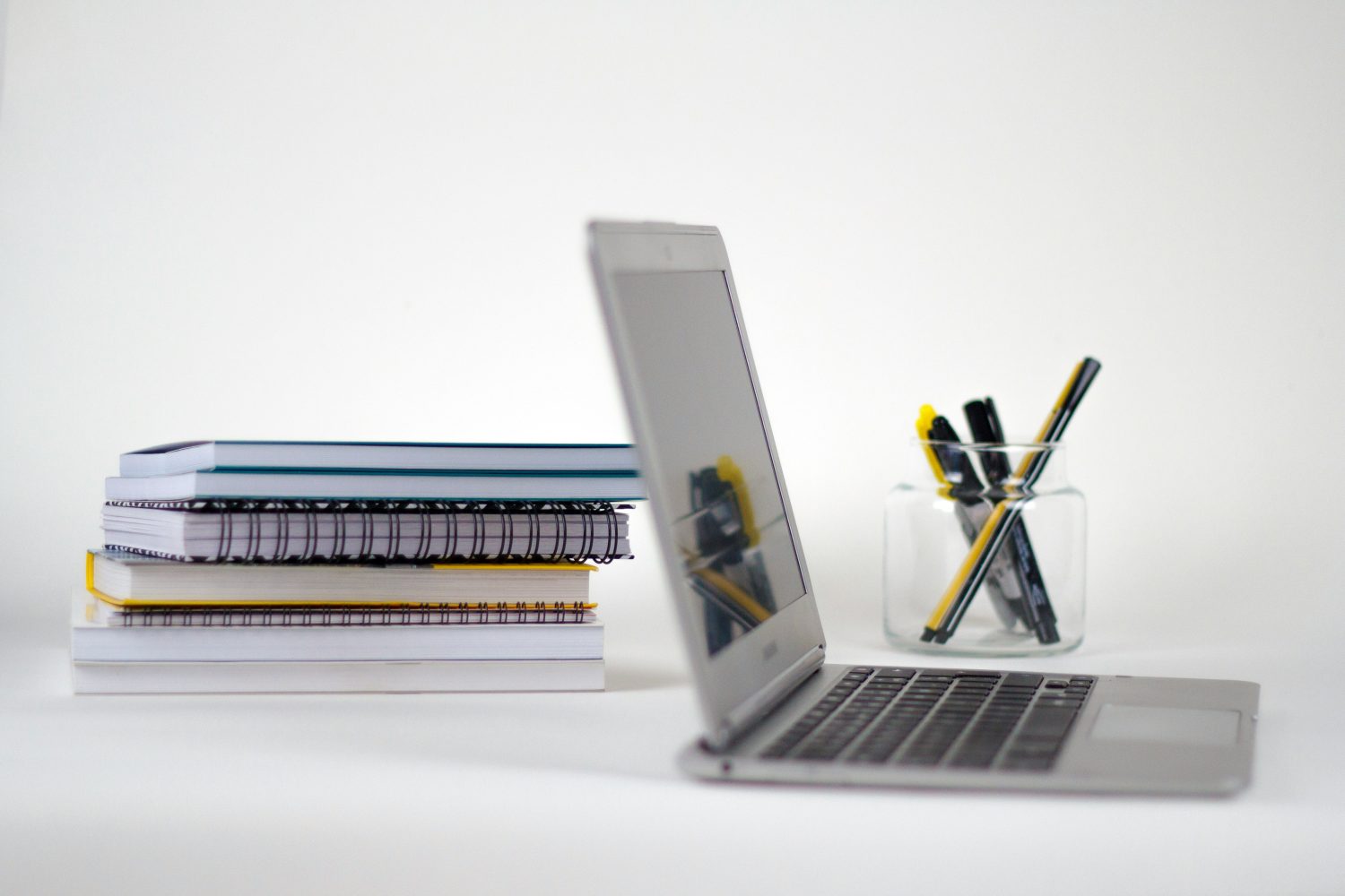 Auf einer weißen Fläche liegt ein aufgeklappter Laptop neben einem Stapel Notizbücher und einem durchsichtigen Behälter mit Stiften und Textmarkern, alles vor einem schlichten weißen Hintergrund – eine ideale Kulisse für die Ausbildung oder den Einsatz in jedem modernen Lehrbetrieb.