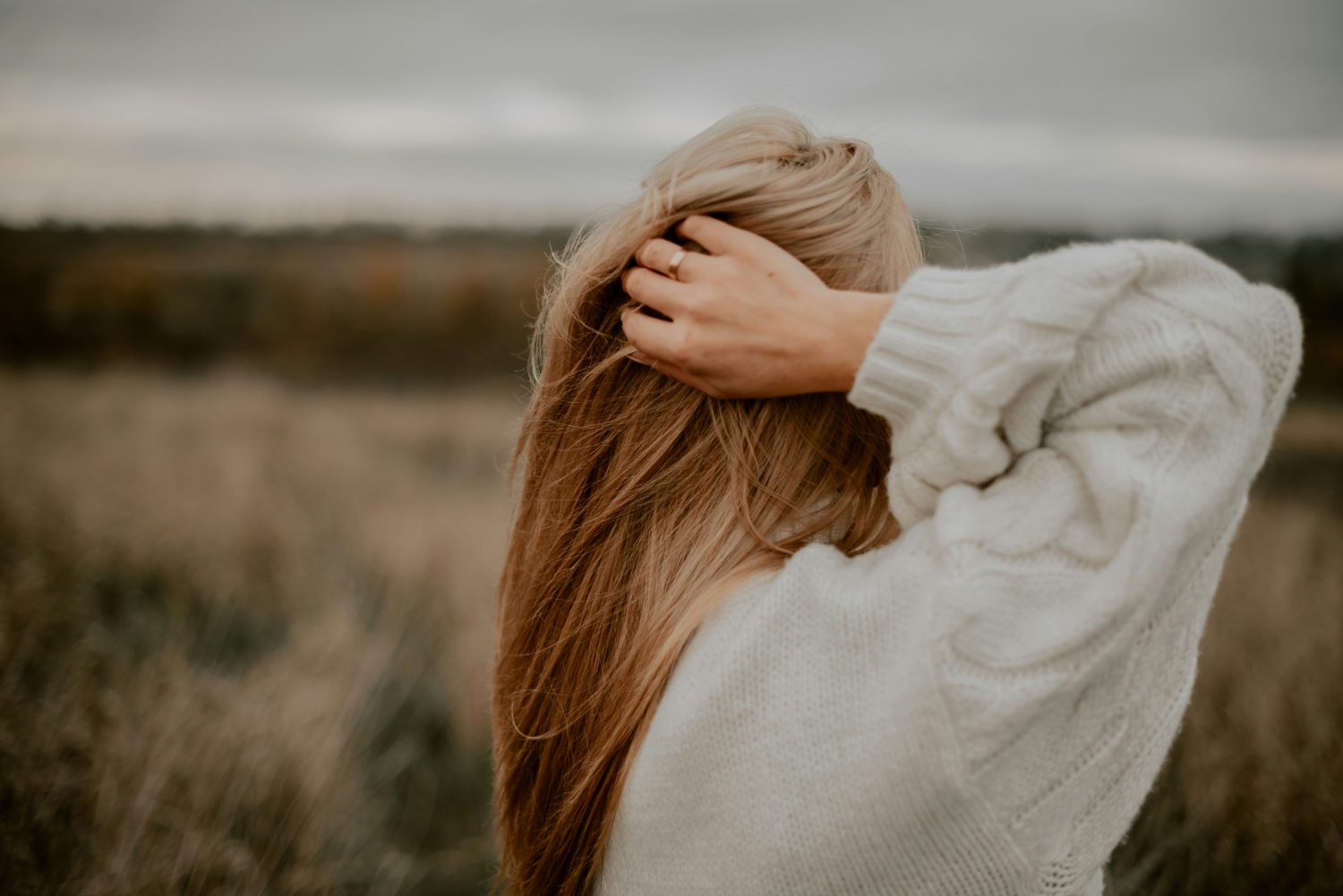 Eine Person mit langem, hellbraunem Haar und einem cremefarbenen Pullover steht in einem Feld, von der Kamera abgewandt, und strahlt in ihrer entspannten Haltung eine kollagene Vitalität aus. Der unscharfe Hintergrund ist herbstlich.
