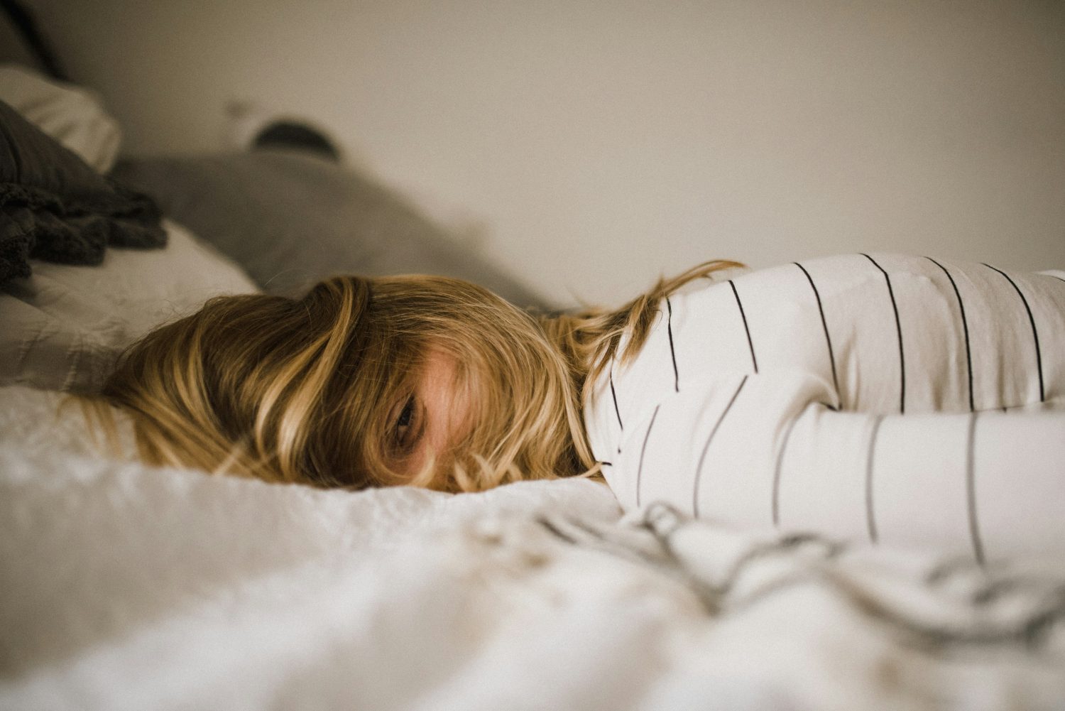 Une personne aux cheveux blonds est allongée face contre le lit et porte une chemise blanche à rayures noires. Son regard, qui reflète peut-être une carence en fer, est tourné vers l'appareil photo. La literie est blanche, avec des oreillers gris en arrière-plan.