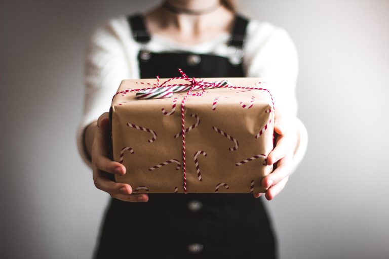 Eine Person in weißem Hemd und schwarzem Overall präsentiert ein Geschenk, das in braunes Papier mit Zuckerstangen-Motiven eingewickelt und mit rot-weißer Kordel verschnürt ist – perfekt für eine kingnature Adventsaktion. Der Hintergrund ist schlicht und unscharf.