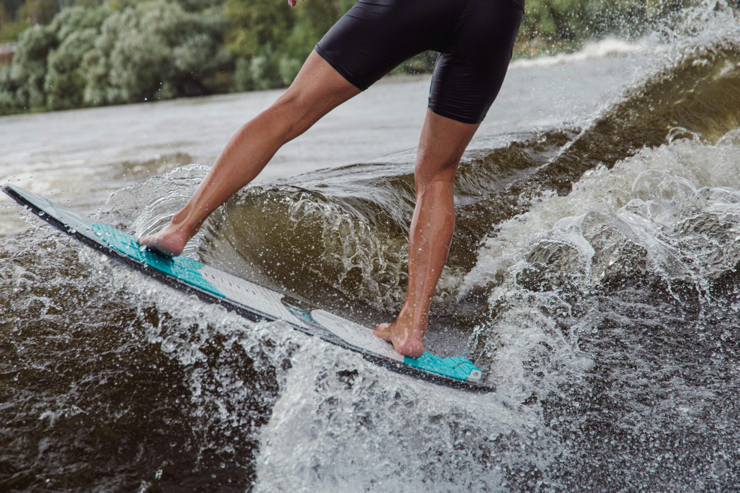 Eine Person im Neoprenanzug reitet auf einem türkis-weißen Surfbrett auf einer Flusswelle, das Wasser plätschert um ihre Beine und im Hintergrund sind Bäume zu sehen - sie fängt die Energie ein, genau wie Mitoflow Vida Produkt Ihre Mitochondrien unterstützt.