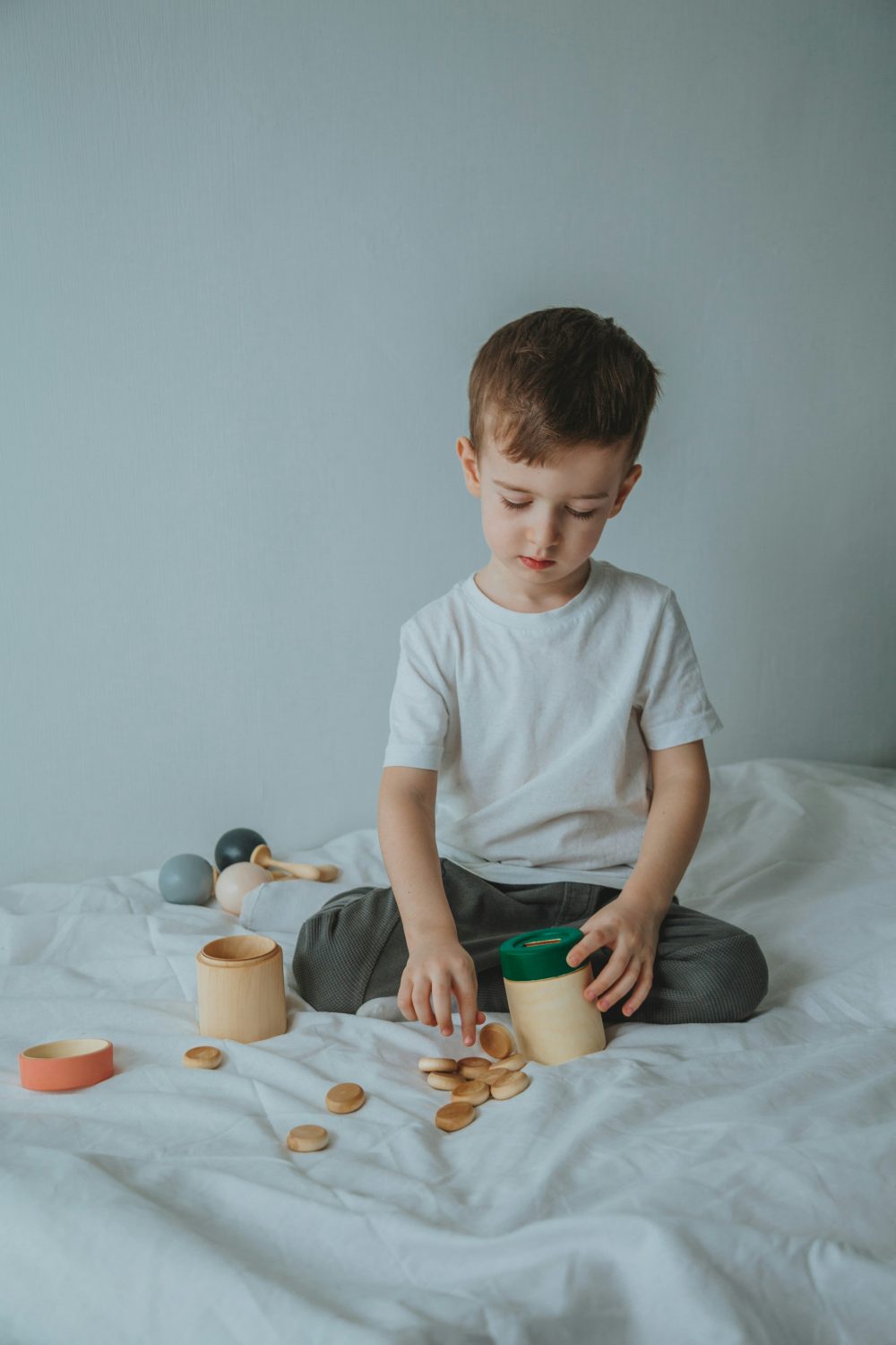 Ein kleiner Junge in weißem T-Shirt und grauer Hose sitzt auf einem weißen Bett, spielt mit Holzspielzeug und sortiert Holzscheiben in bunte Behälter – eine beruhigende Beschäftigung, die Kinder mit Autismus-Spektrum-Störungen oft genießen. Der Hintergrund ist schlicht und minimalistisch.