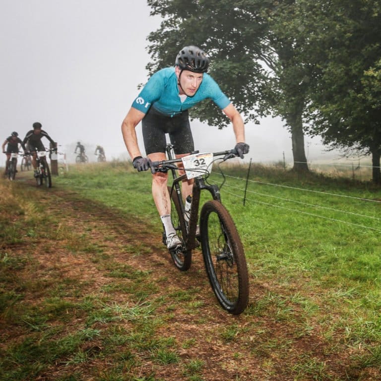 Mountainbiker Johannes Közle, bekleidet mit einem blauen Trikot und Helm, fährt während eines Rennens mit seinem Mountainbike auf einem grasbewachsenen, schlammigen Pfad. Andere Radfahrer folgen ihm, und im nebligen Hintergrund sind Bäume zu sehen.