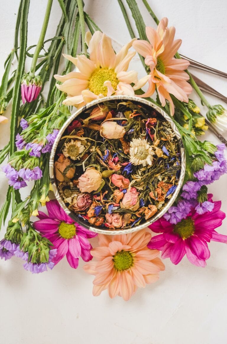 Eine Dose mit losem, getrocknetem Kräutertee mit bunten Blütenblättern und Kräutern steht umgeben von frischen Gänseblümchen und anderen leuchtenden Blumen auf einer weißen Oberfläche und bietet die beste Naturkosmetik für deine Haut.