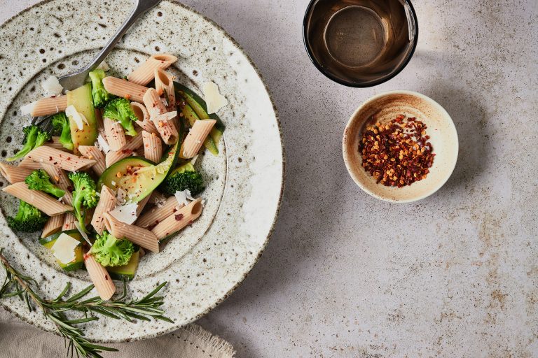 Ein Teller Penne mit Brokkoli und Zuckerschoten, garniert mit Käse und Chiliflocken, steht neben einer Schüssel mit Chiliflocken und einem Glas Wasser auf einer gesprenkelten Fläche. Ein Rosmarinzweig daneben sorgt für aromatischen Geschmack.
