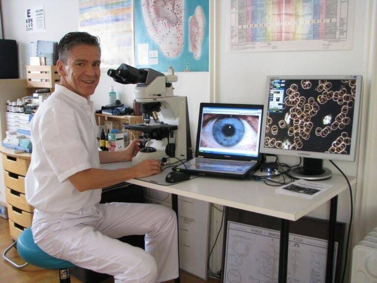 Ein Mann in weißer Uniform sitzt an einem Schreibtisch mit einem Mikroskop. Zwei Monitore zeigen Nahaufnahmen eines Auges und kreisförmiger Zellen. Im Hintergrund sind wissenschaftliche Diagramme und Geräte in Olivier Struchens Naturheilpraxis zu sehen.
