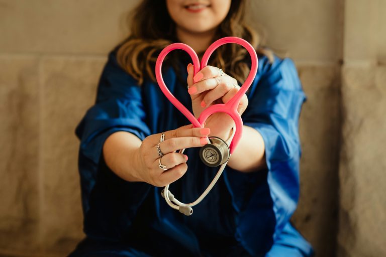 Eine Frau in blauem Outfit hält ein rosa herzförmiges Stethoskop und lächelt leicht. Der Fokus auf ihre Hände und ihr Stethoskop unterstreicht die Fürsorge und das Mitgefühl im Gesundheitswesen – eine inspirierende Erinnerung, gerade wenn unsere Stimme gefragt ist.